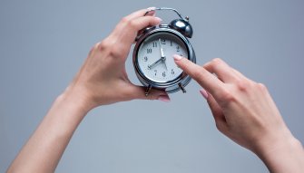 A silver alarm clock held by a person's hands with a gray background.